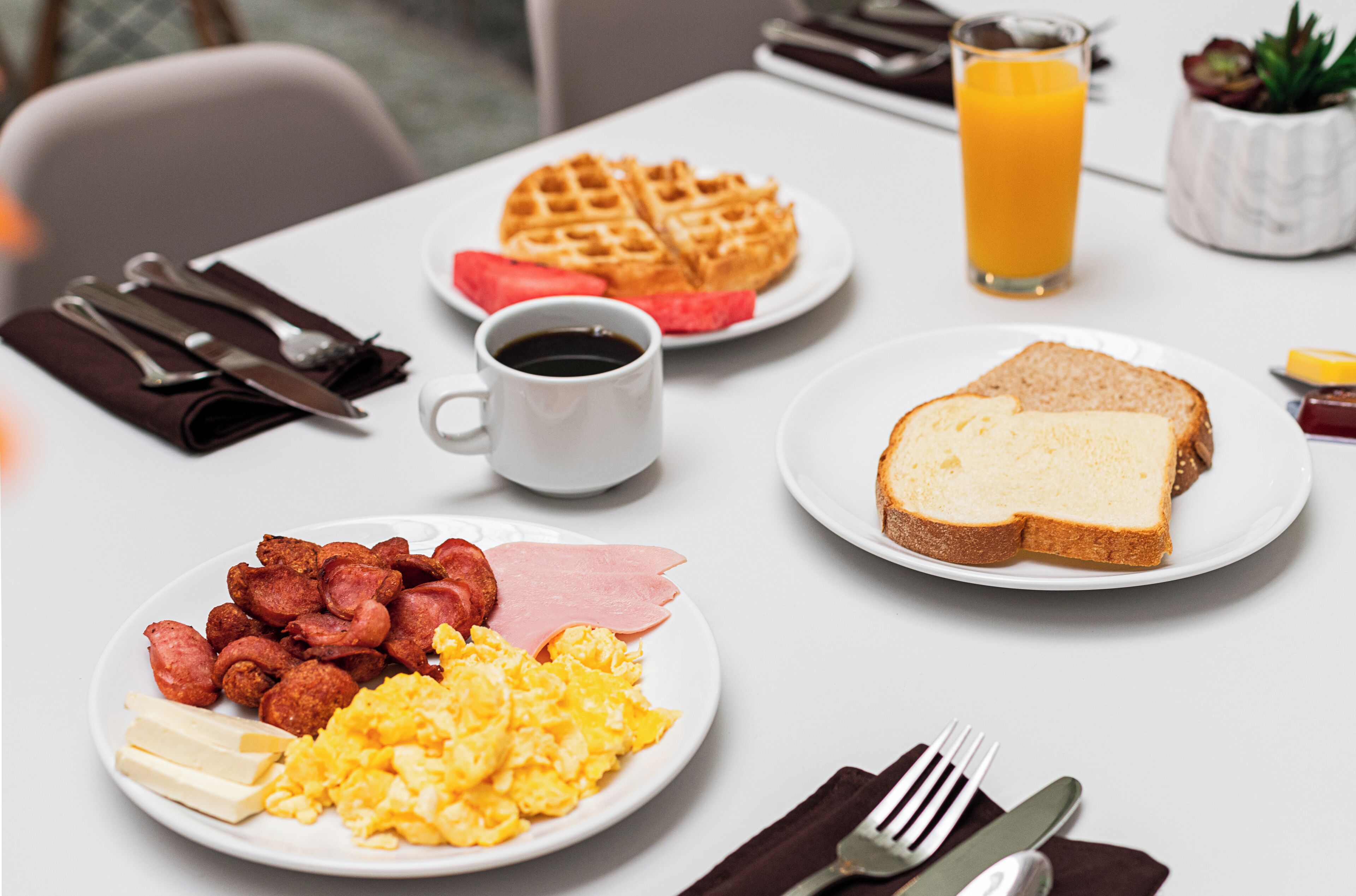 Une table de petit-déjeuner avec des assiettes contenant des œufs brouillés, du bacon, des gaufres, des toasts et un verre de jus d’orange.