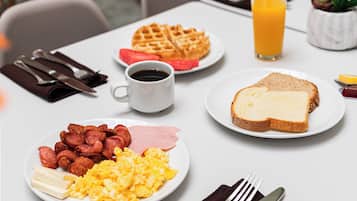 Une table de petit-déjeuner avec des assiettes contenant des œufs brouillés, du bacon, des gaufres, des toasts et un verre de jus d’orange.