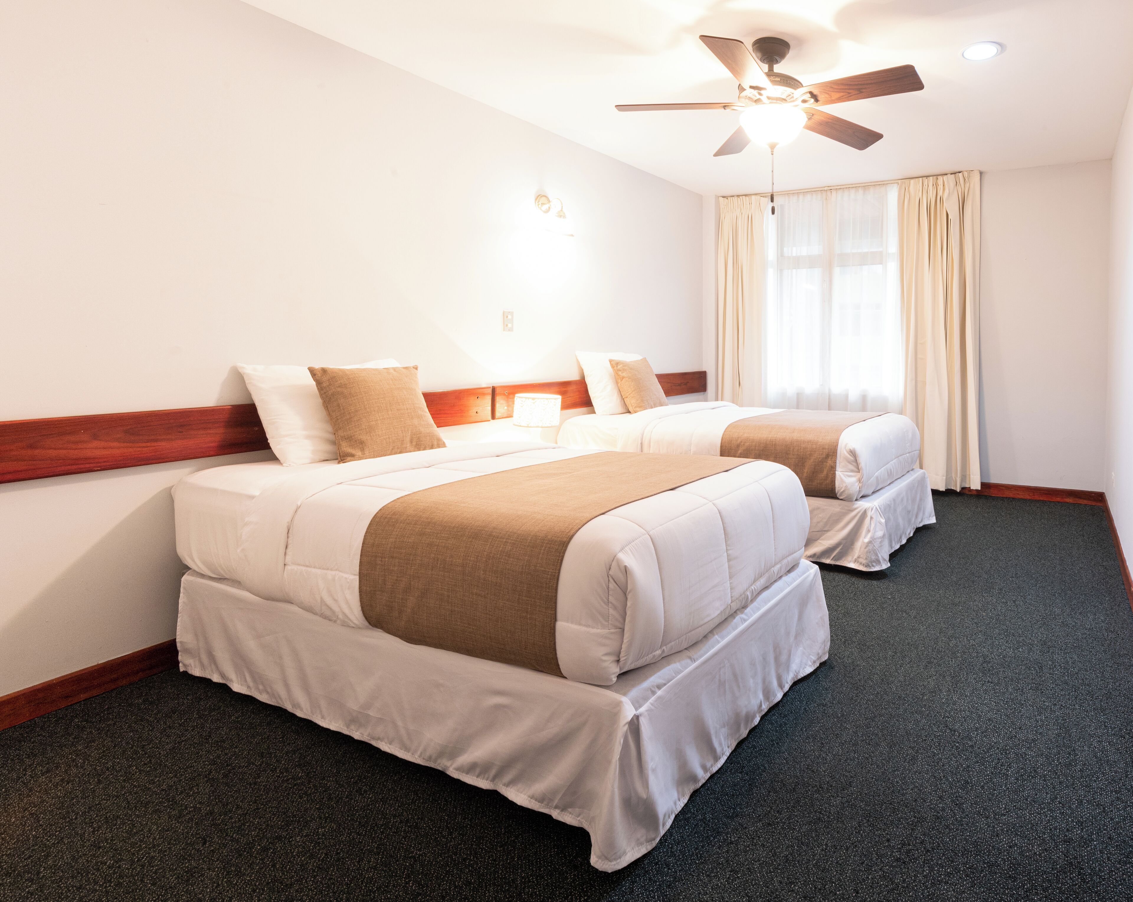 A hotel room with two beds, a ceiling fan, a window with curtains, and a carpeted floor.