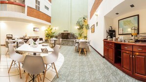 A hotel dining area with multiple tables and chairs, a kitchen area with wooden cabinets, and a stone fireplace.
