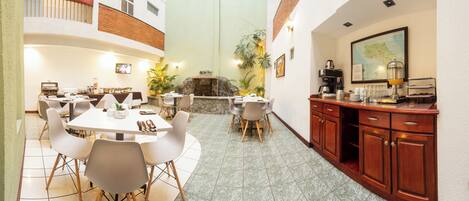 A hotel dining area with multiple tables and chairs, a kitchen area with wooden cabinets, and a stone fireplace.