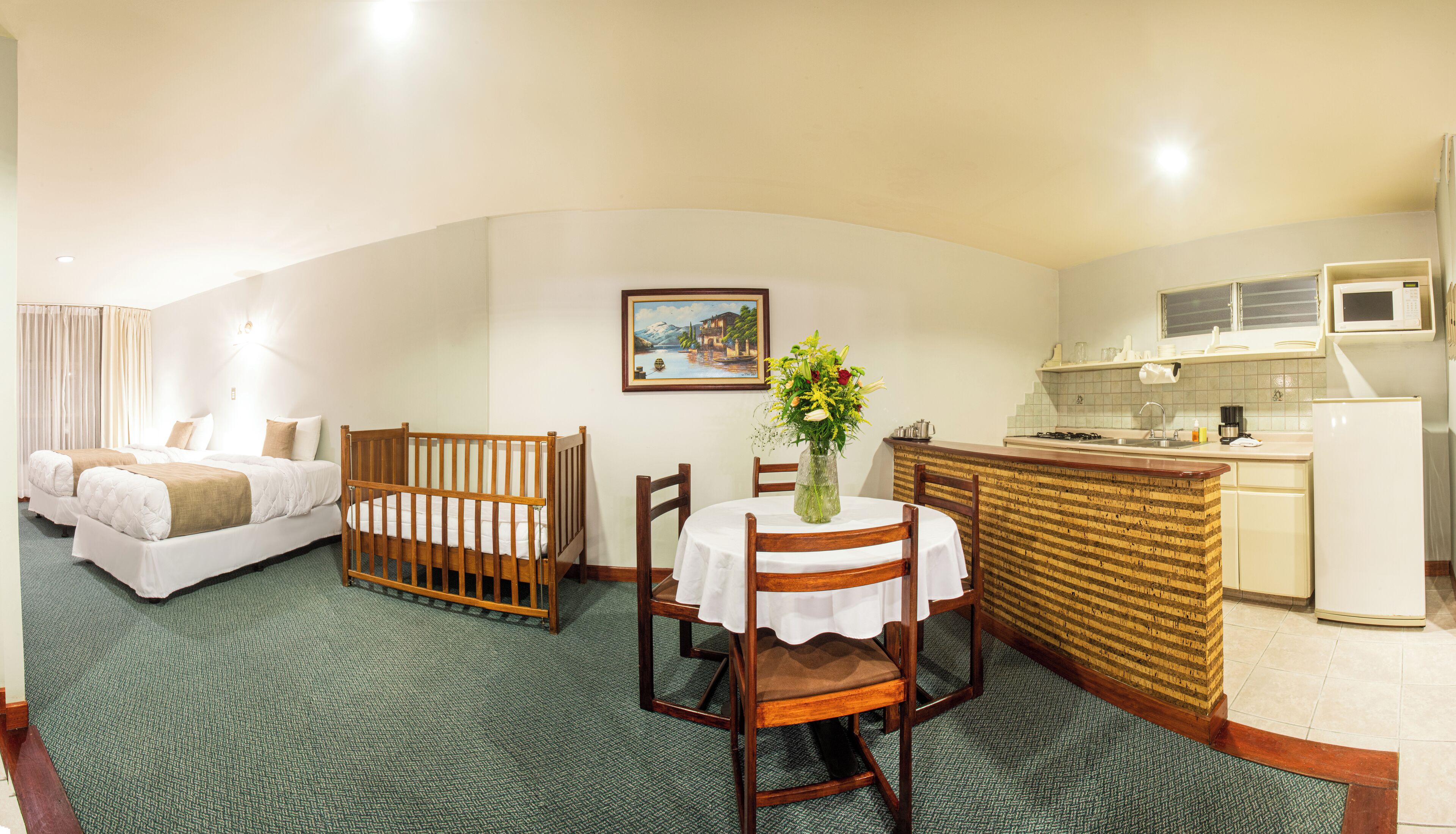 A room with a bed, a crib, a dining table with chairs, a kitchen area, and a painting on the wall.