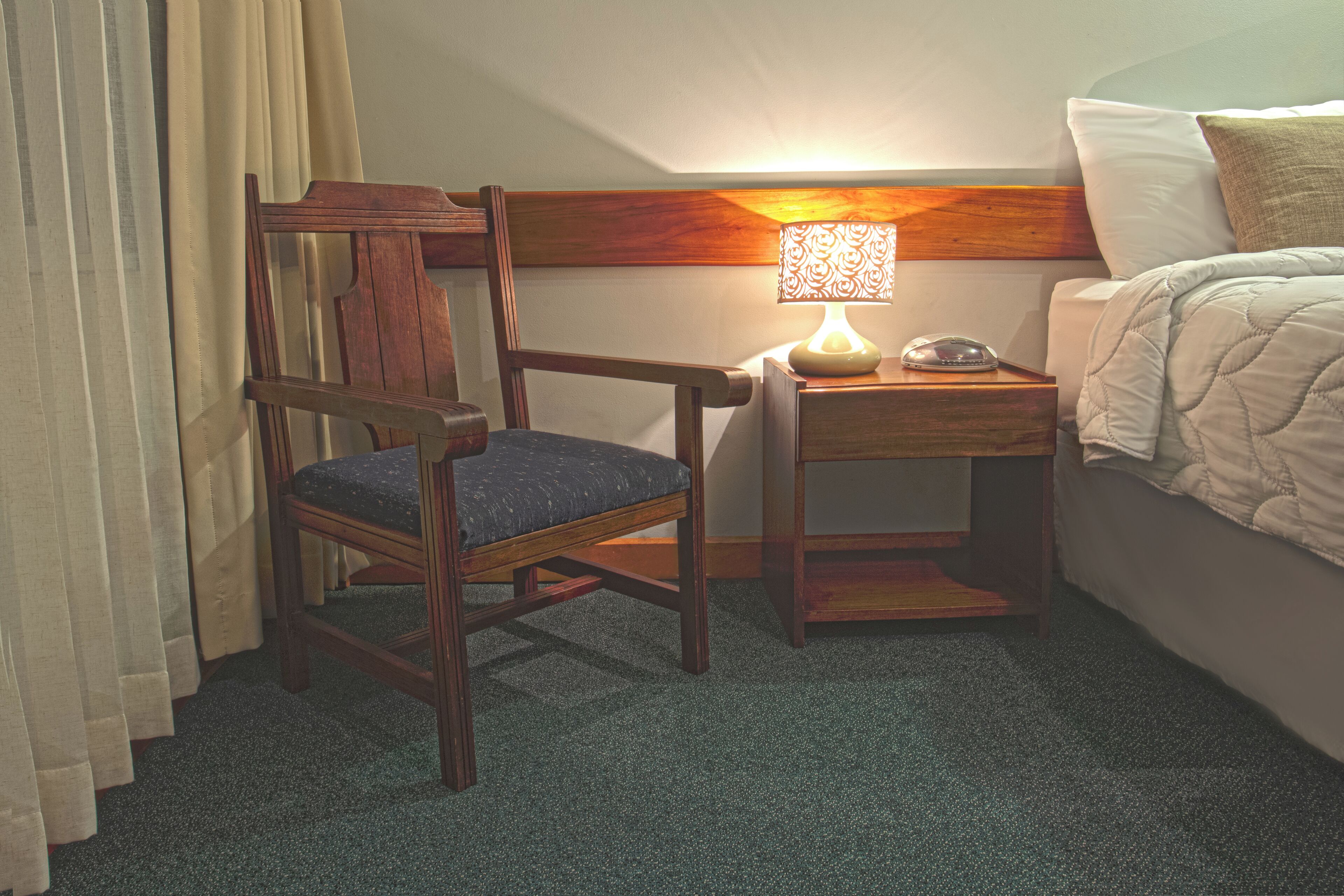 Une chambre d’hôtel avec une chaise en bois, une table de chevet avec une lampe et un lit recouvert d’une courtepointe.