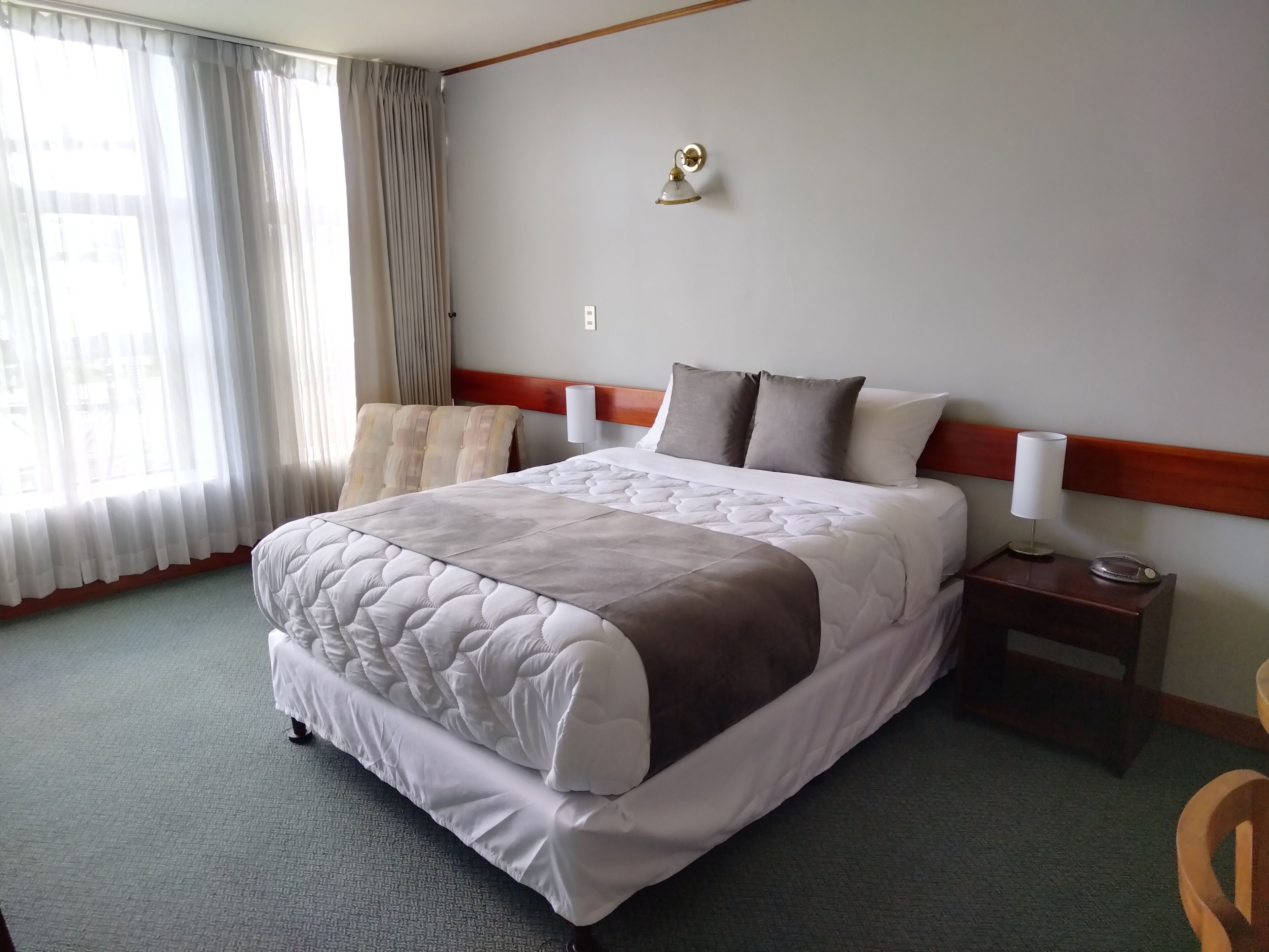 Une chambre d’hôtel avec un grand lit, une tête de lit en bois, une table de chevet avec une lampe et une vue sur la ville à travers de fines rideaux.