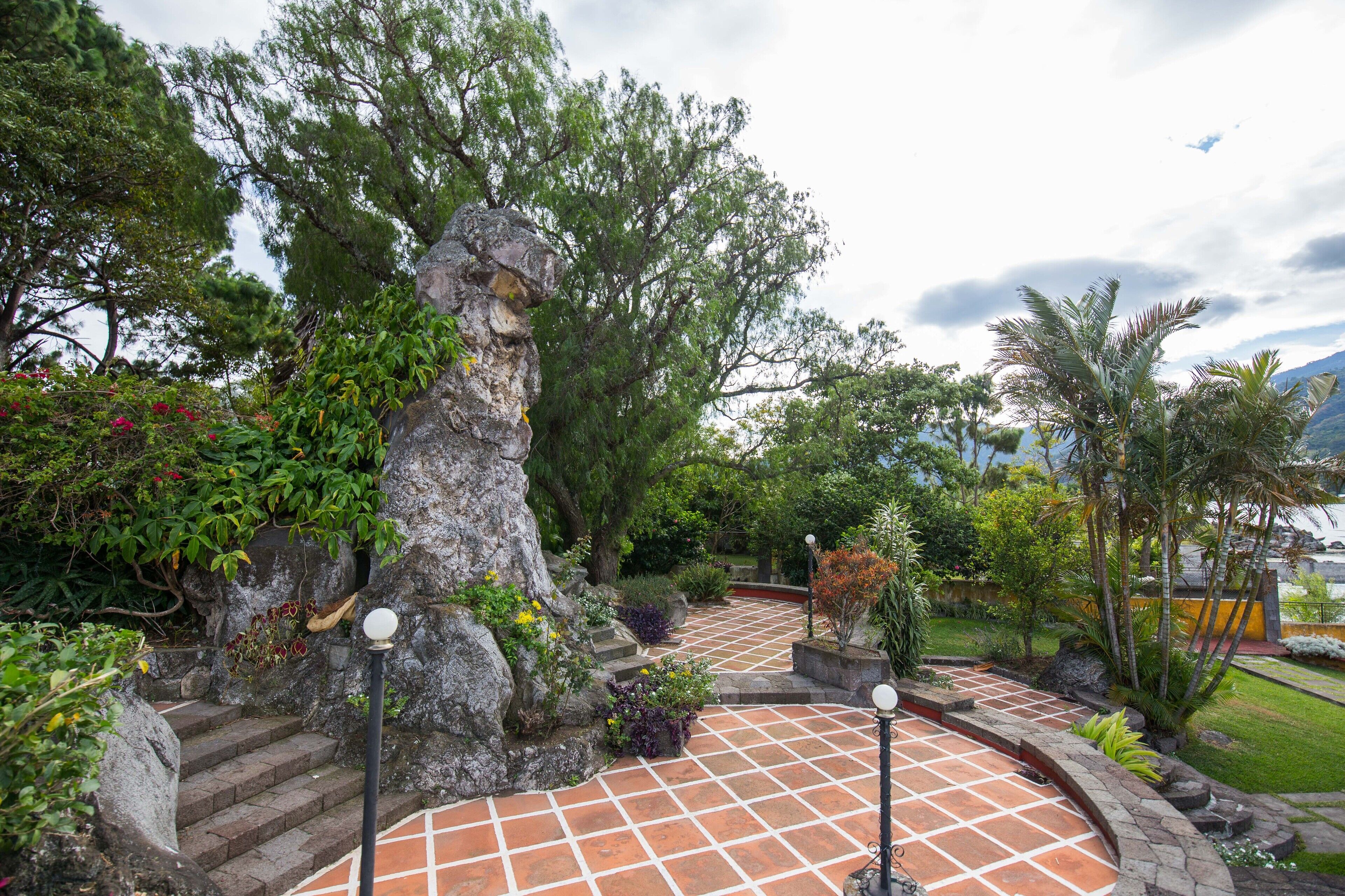 A garden with a stone staircase leading up to a rock formation, surrounded by lush greenery and a tiled pathway.