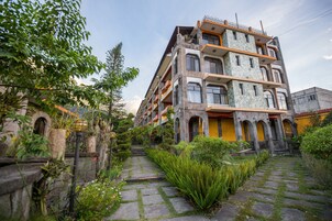 Un edificio de varios pisos con balcones y fachada de piedra, rodeado de vegetación y un camino de piedra.