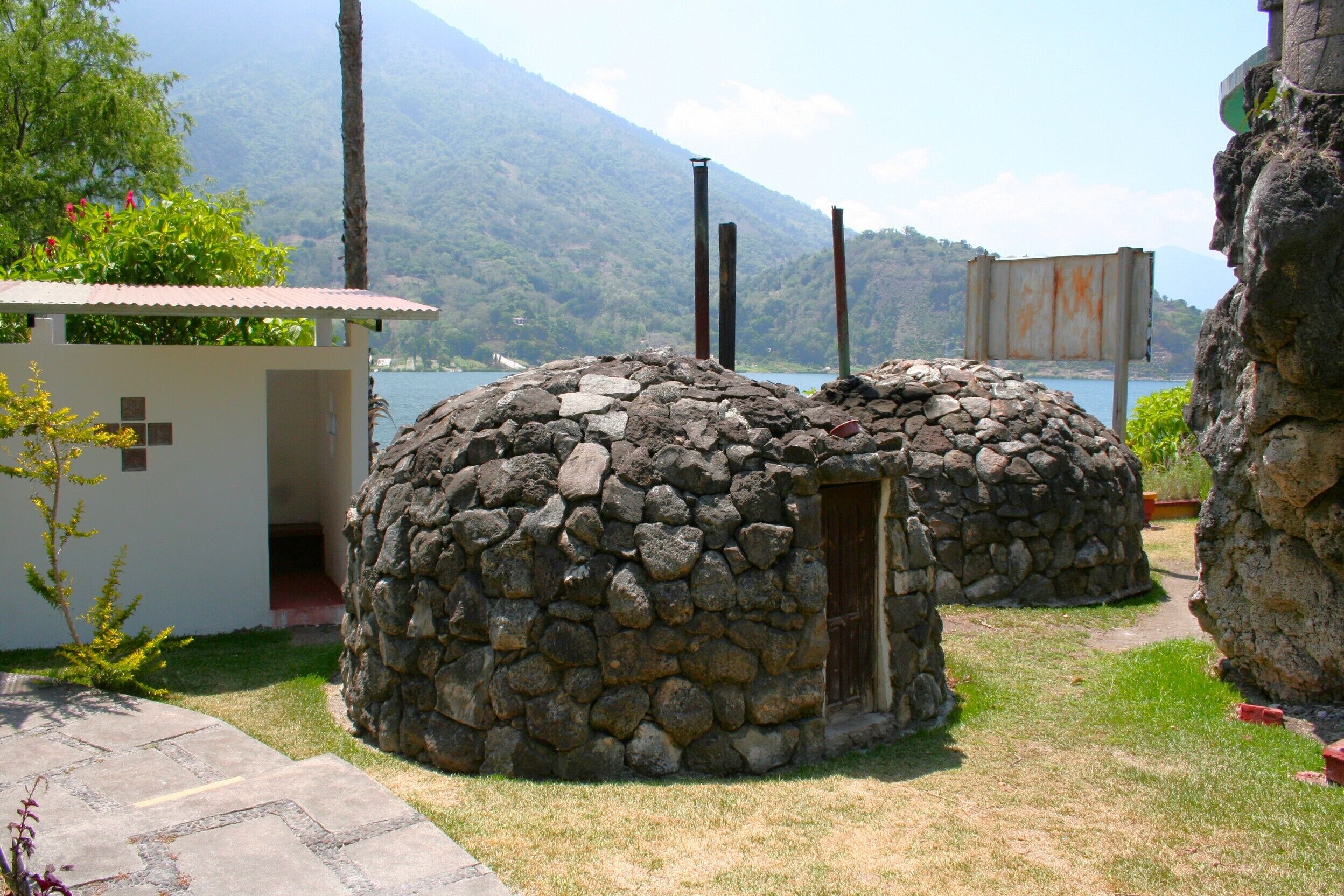 Two stone structures with a small door, surrounded by grass and a pathway, near a body of water and mountains.