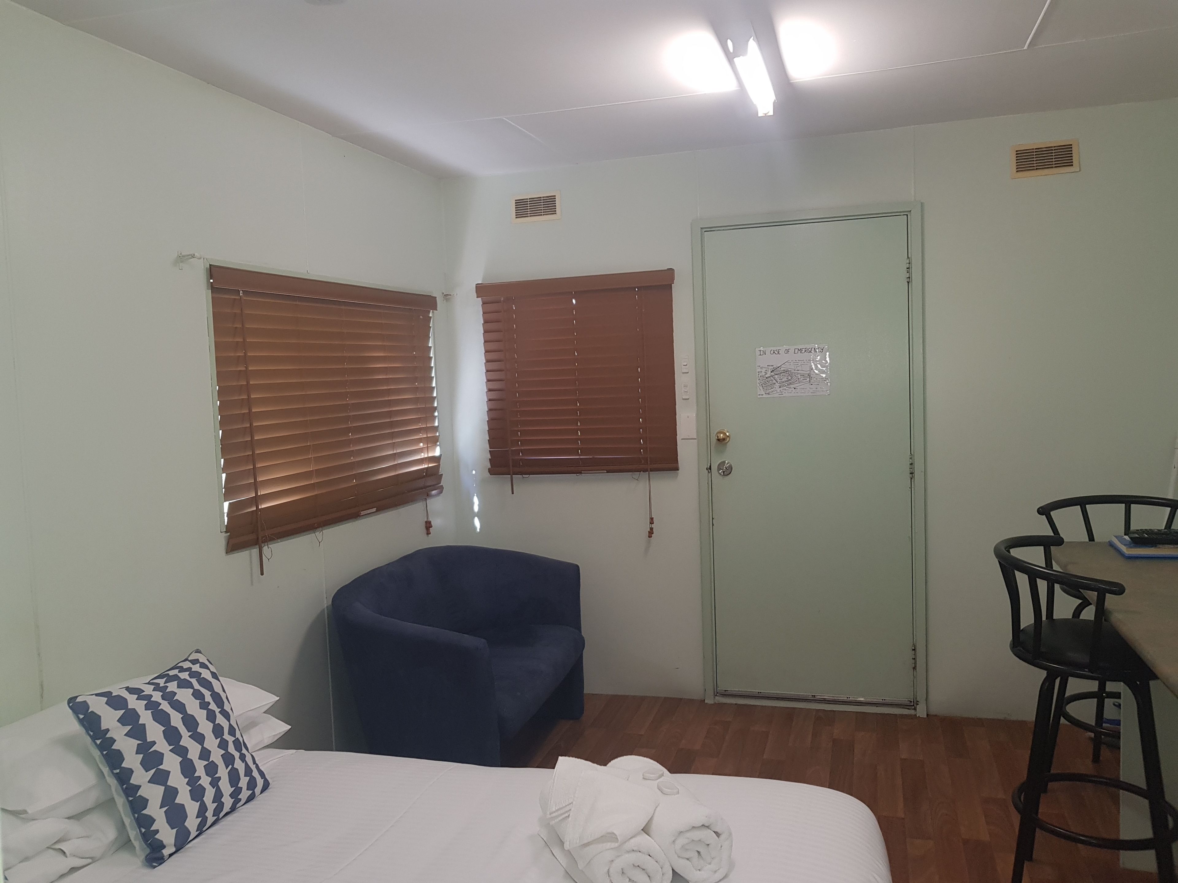 A room with a bed, a blue chair, a desk with a chair, and a door with an emergency exit sign.