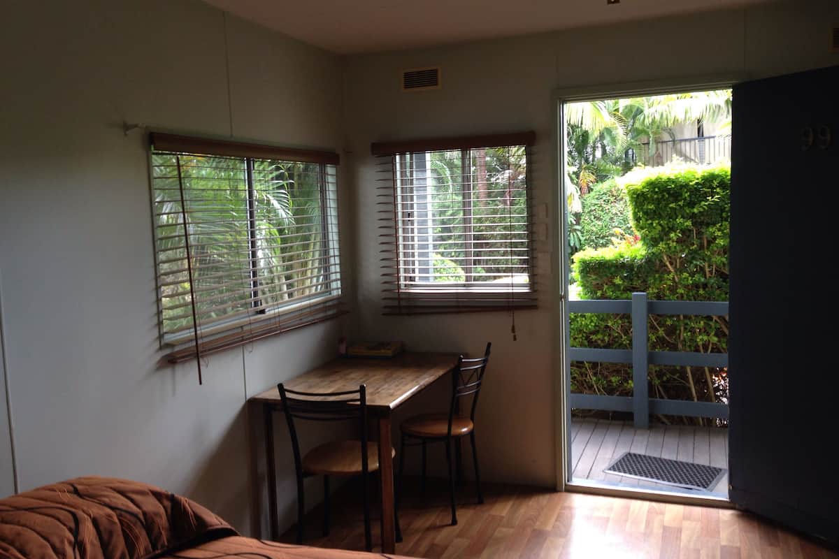 A room with a wooden table and chairs, a bed, and a sliding glass door leading to an outdoor area with a blue fence and greenery.