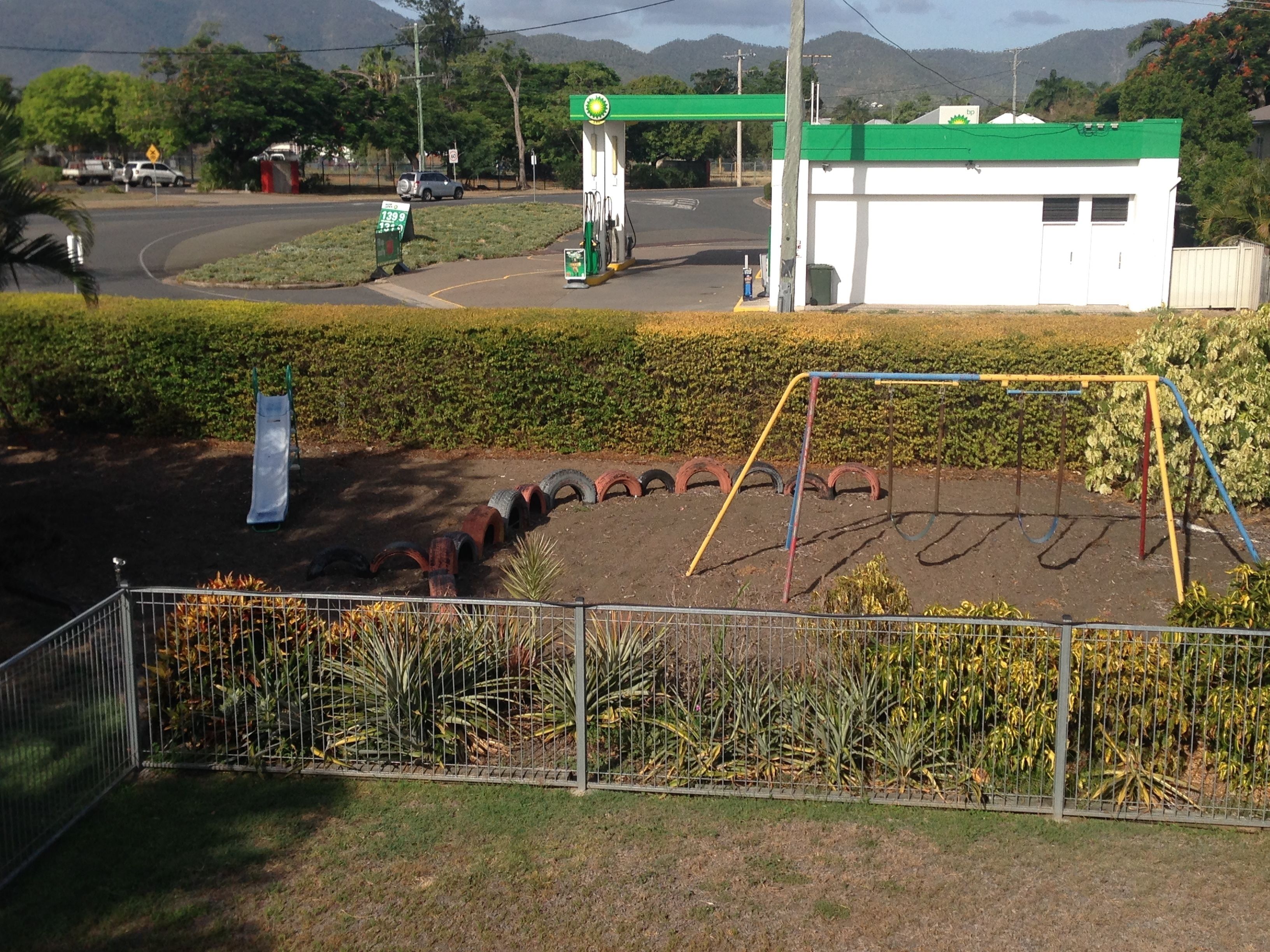 A gas station with a green canopy, a playground with a swing set, and a fenced area with plants.