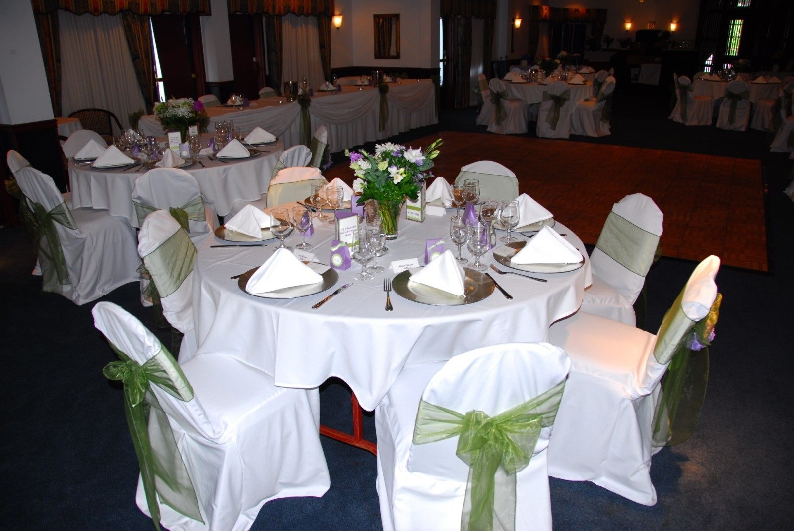 Une salle de banquet avec des tables rondes dressées pour un événement formel, chacune étant recouverte d’une nappe blanche et agrémentée de couverts en argent et de verres.