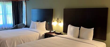 A hotel room with two beds, a nightstand with a lamp, and a window with curtains.