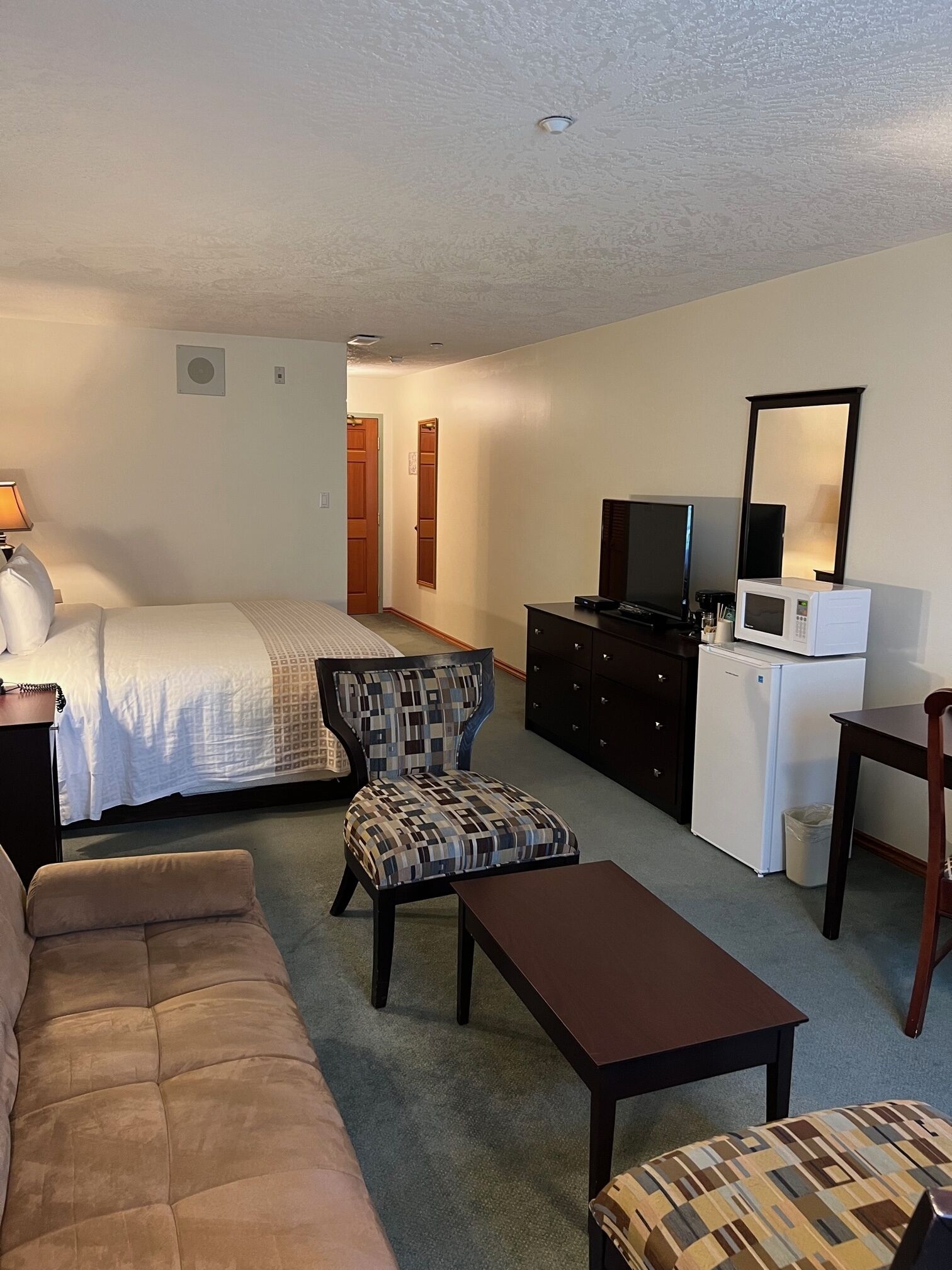 A hotel room with a bed, a sofa, a chair, a table, a dresser, a television, and a microwave.