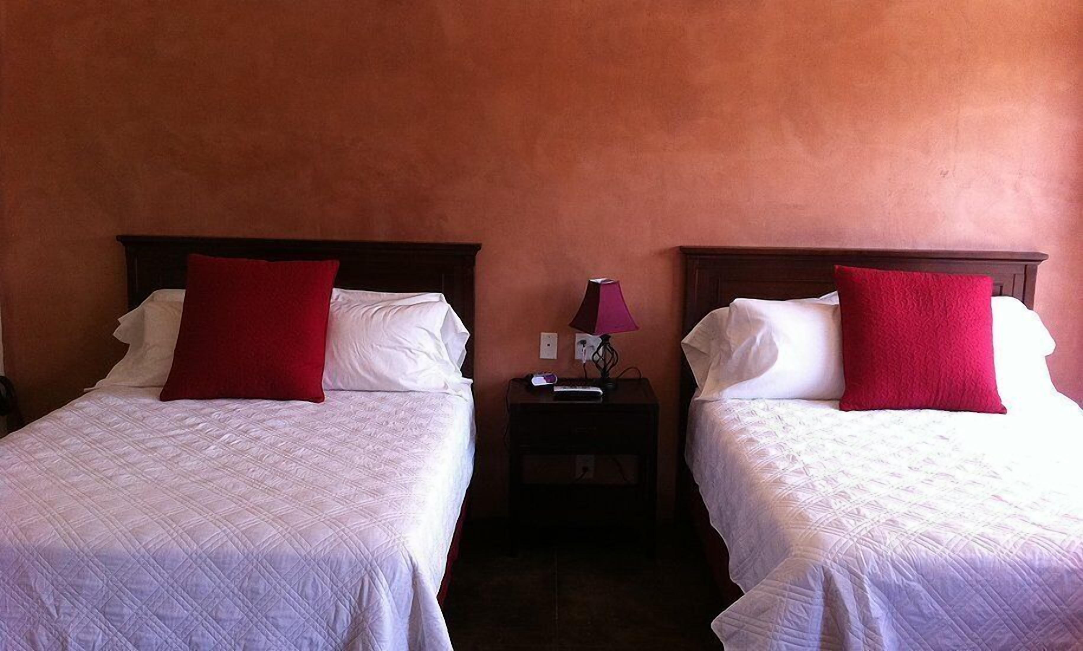 Two beds with white linens and red pillows, a nightstand with a lamp, and a wall with a textured finish.
