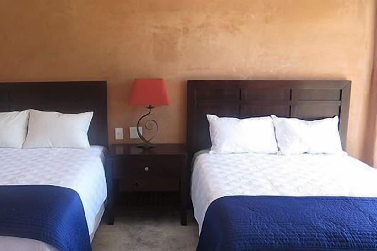 Dos camas con sábanas blancas y colchas azules, una mesita de noche de madera con una lámpara roja, y una pared naranja con textura.