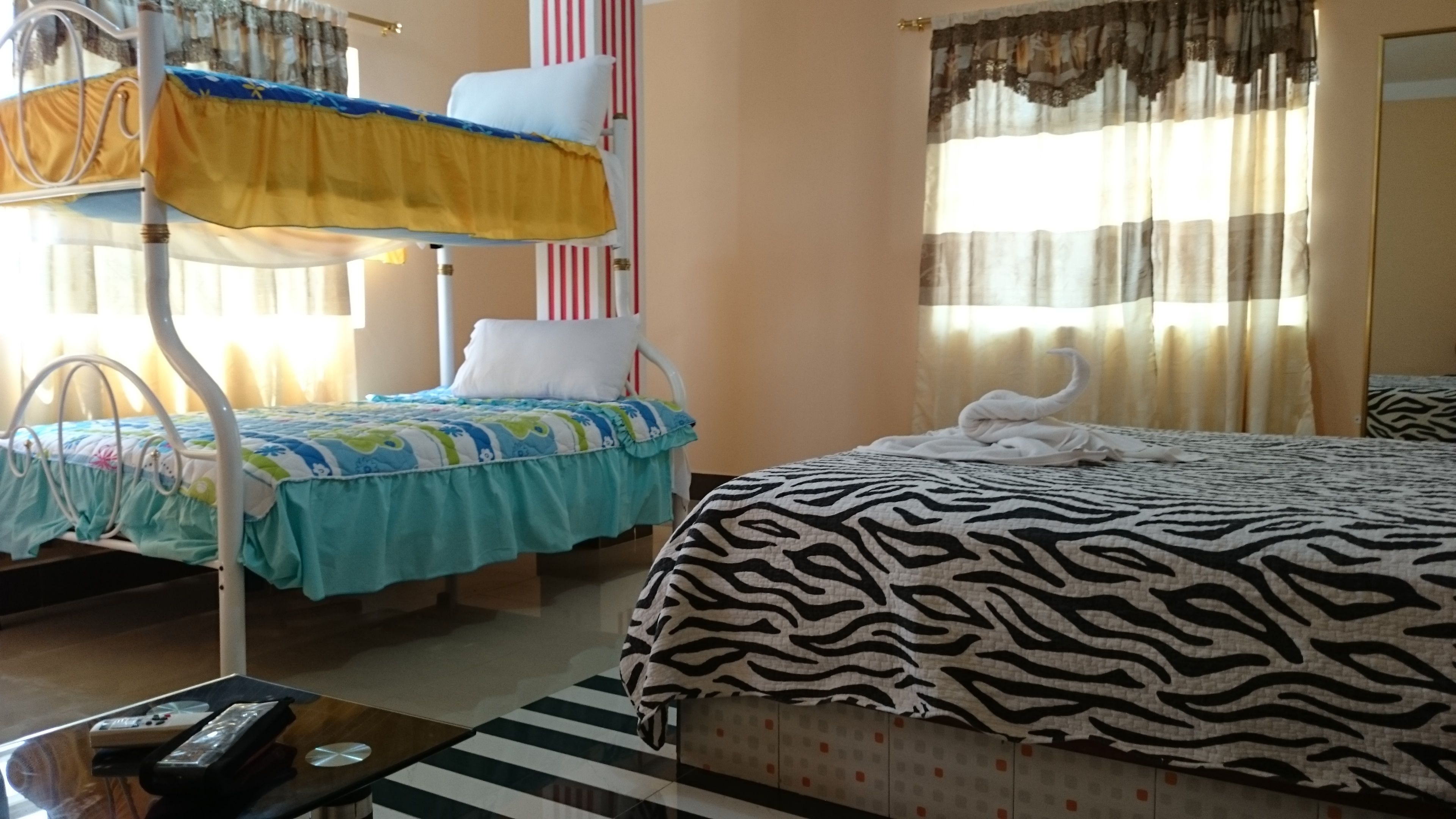 A hotel room with a bunk bed, a single bed with a zebra-patterned bedspread, a small table with a remote control, and a window with curtains.
