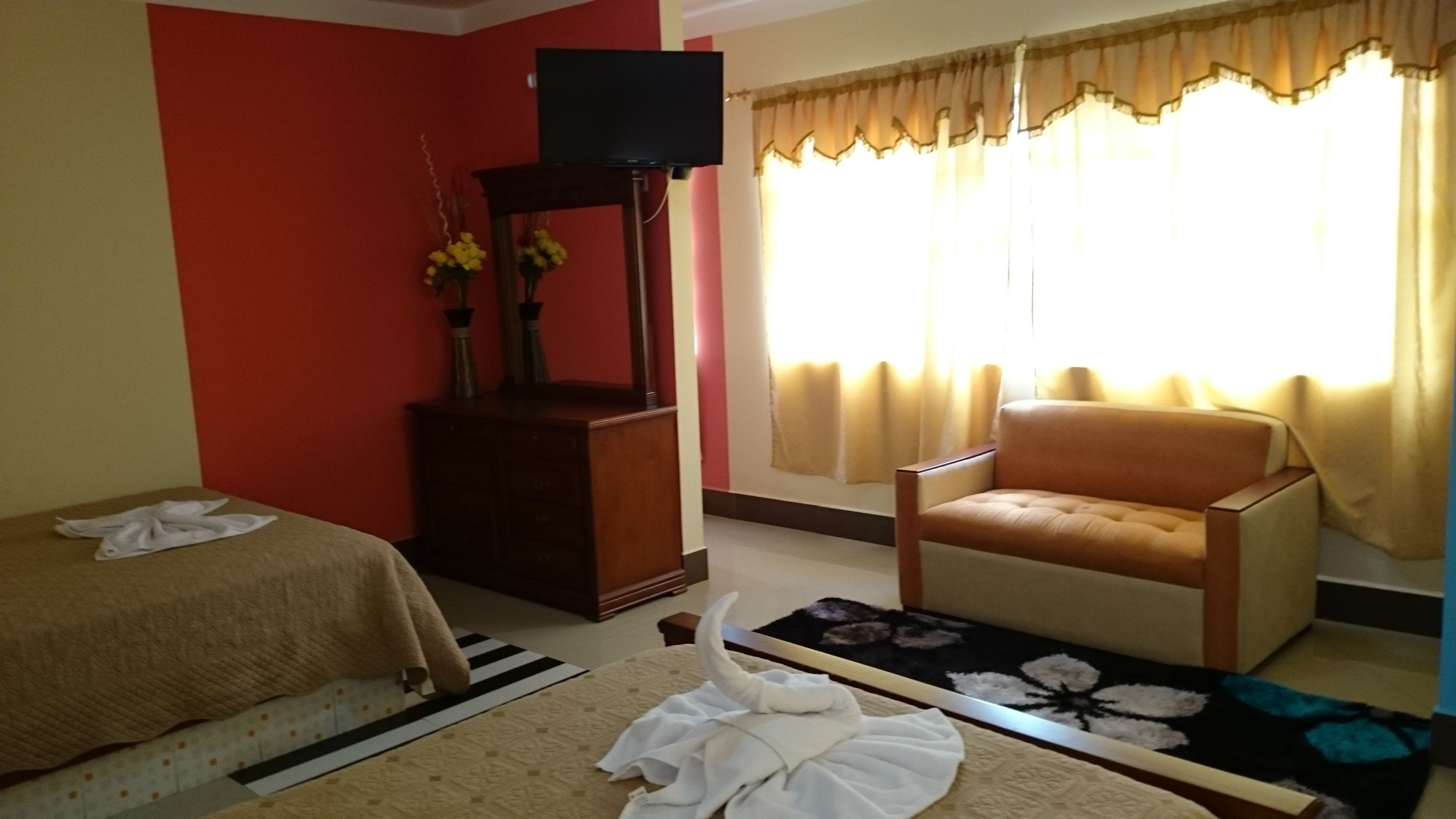 Una habitación de hotel con cama, tocador de madera, televisor, sillón y ventana con cortinas.