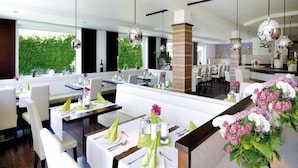 A modern restaurant with white tables and chairs, hanging pendant lights, and a view of greenery outside. - Pension Bruderhofer (ALTMUENSTER)
