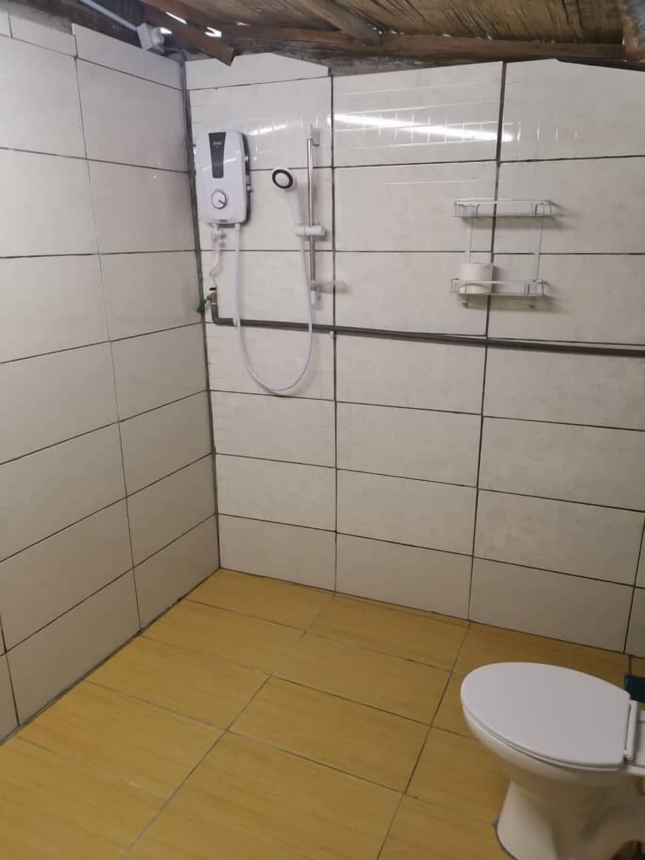 A small bathroom with white tiles, a toilet, a shower, and a shelf with toiletries.