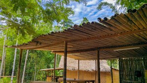 An outdoor seating area with wooden tables and chairs, a thatched-roof structure, and bamboo poles.