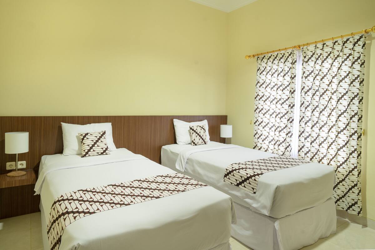 Habitación de hotel con dos camas, cada una con cubrecama y almohadas estampadas, cabeceros de madera, lámparas de noche y una ventana con cortinas estampadas.