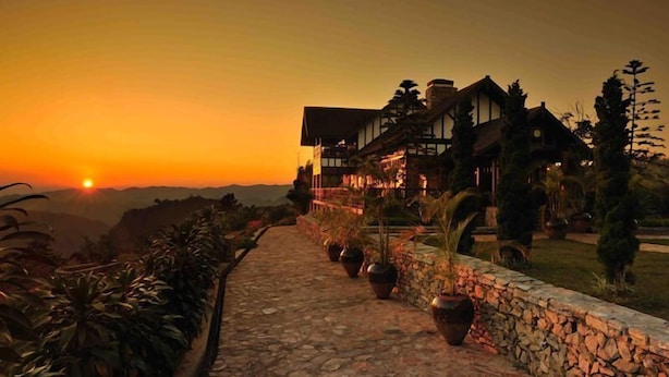 Un chemin de pierre mène à un grand bâtiment à plusieurs étages, de style architectural traditionnel, entouré d’une végétation luxuriante et offrant une vue sur un paysage montagneux au coucher du soleil.