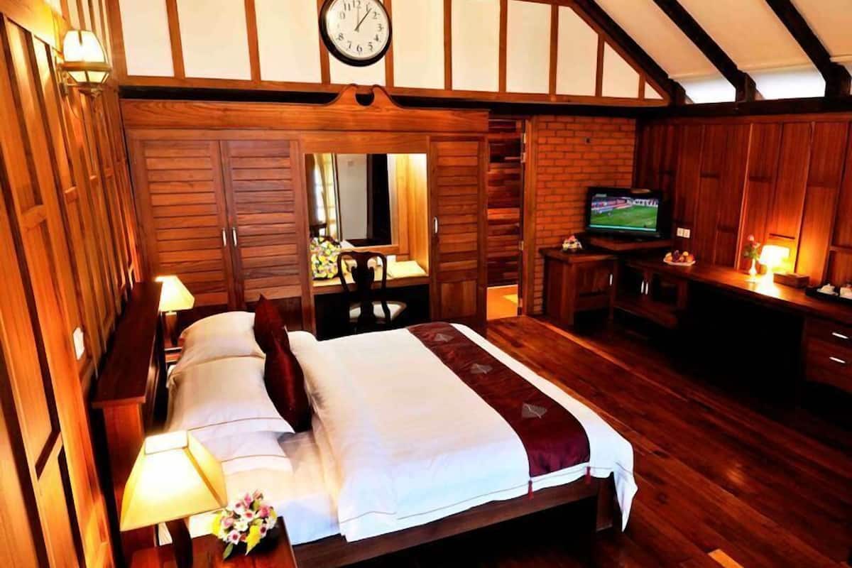 A wooden-floored bedroom with a bed, bedside table, lamp, and a clock on the wall.
