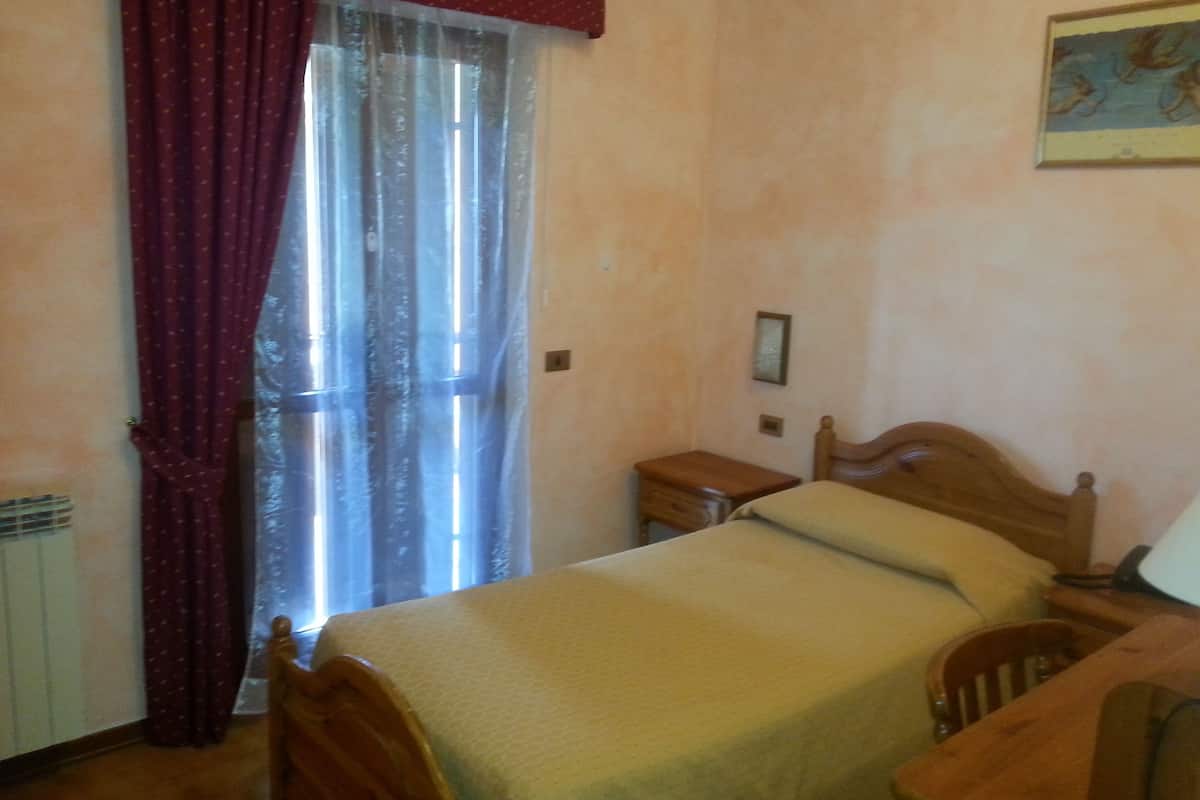 A bedroom with a bed, a wooden nightstand, a chair, a window with curtains, and a wall-mounted picture.