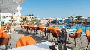 An outdoor dining area with orange chairs and white tables, overlooking a pool with umbrellas and palm trees. - Bouradanis (Kos)