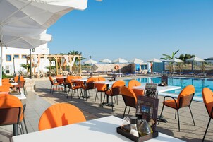 Un área de comedor al aire libre con sillas anaranjadas y mesas blancas, con vistas a una piscina con sombrillas y palmeras.