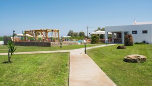 Un sendero conduce a una zona de piscina con una pérgola de madera y sombrillas, un edificio blanco con amplios ventanales y un muro de piedra.