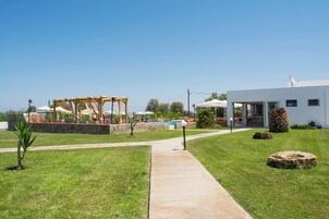 Un sendero conduce a una zona de piscina con una pérgola de madera y sombrillas, un edificio blanco con amplios ventanales y un muro de piedra.