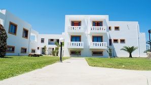 A white building with multiple balconies and windows, surrounded by a well-maintained lawn and a clear blue sky. - Bouradanis (Kos)