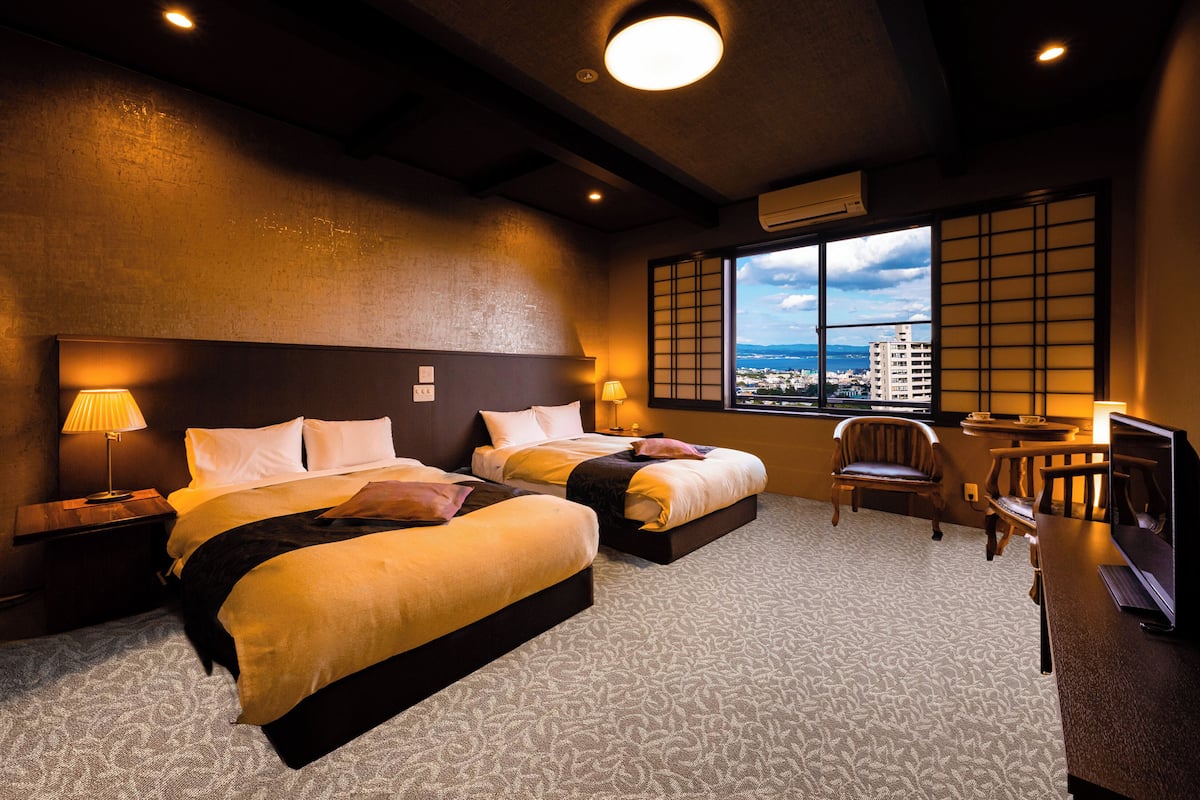 A hotel room with two beds, a desk, a chair, and a view of the city through large windows.
