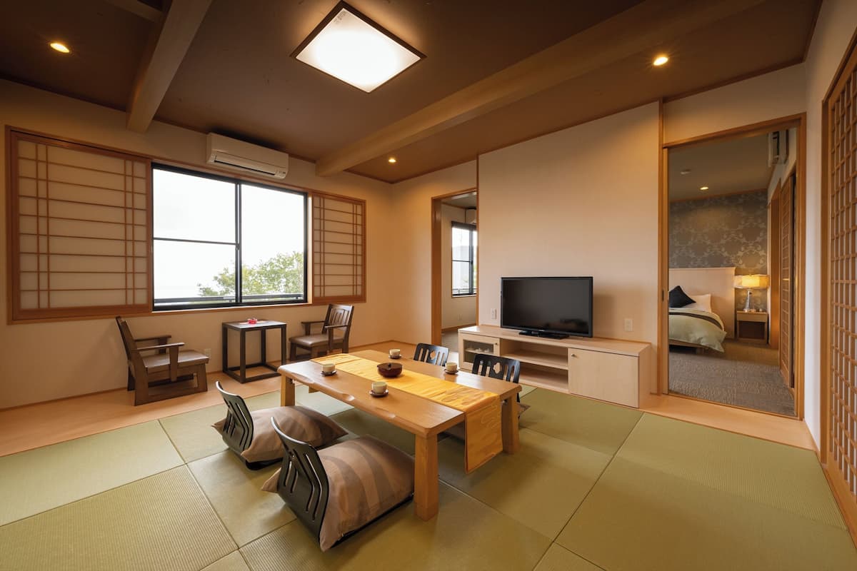 A traditional Japanese-style room with tatami flooring, a low table with cushions, a television, and sliding doors leading to a bedroom.