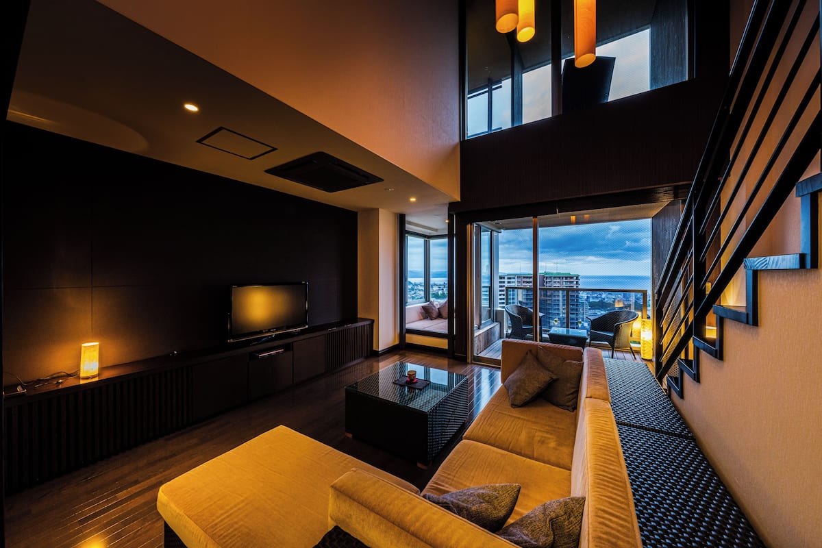 A modern living room with a sofa, a coffee table, and a TV. There is a balcony with a view of the city.