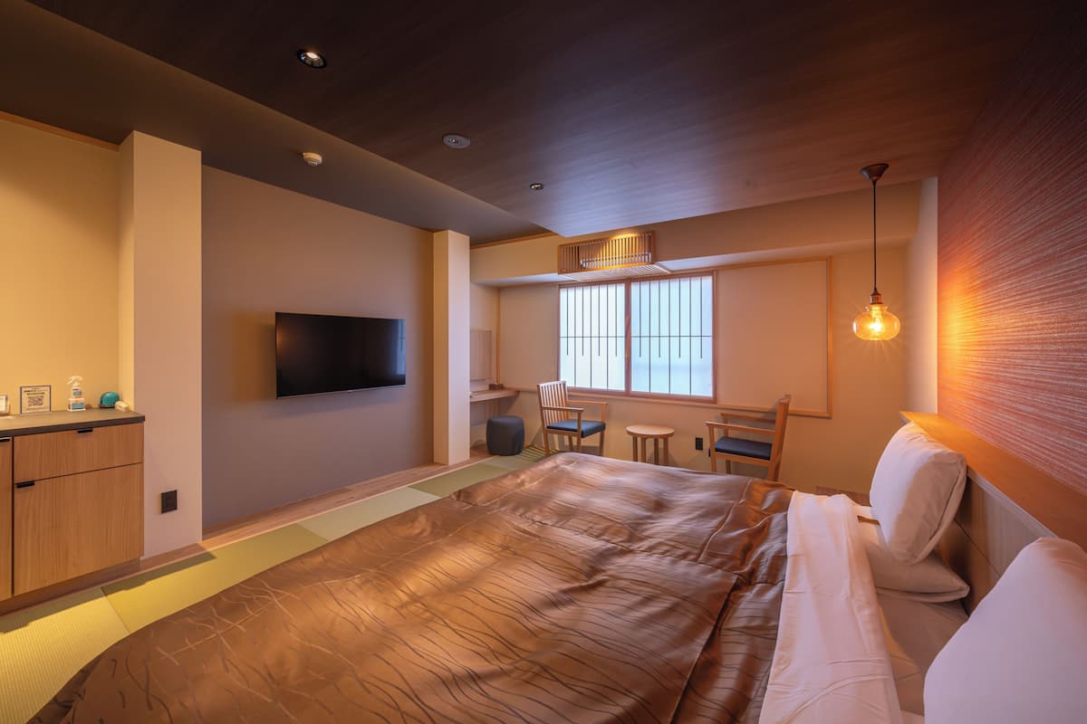 A modern hotel room with a large bed, a flat-screen TV mounted on the wall, a small seating area with a table and chairs, and a window providing natural light.