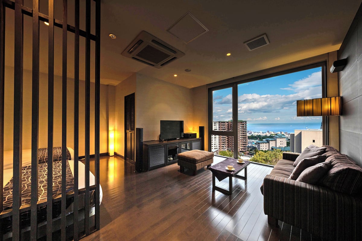 A modern living room with a sofa, ottoman, and a TV on a stand. There is a balcony with a view of the city and the sea.