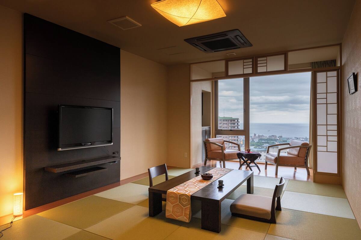 A modern living room with a flat-screen TV, a dining table with chairs, and a view of the city through large windows.