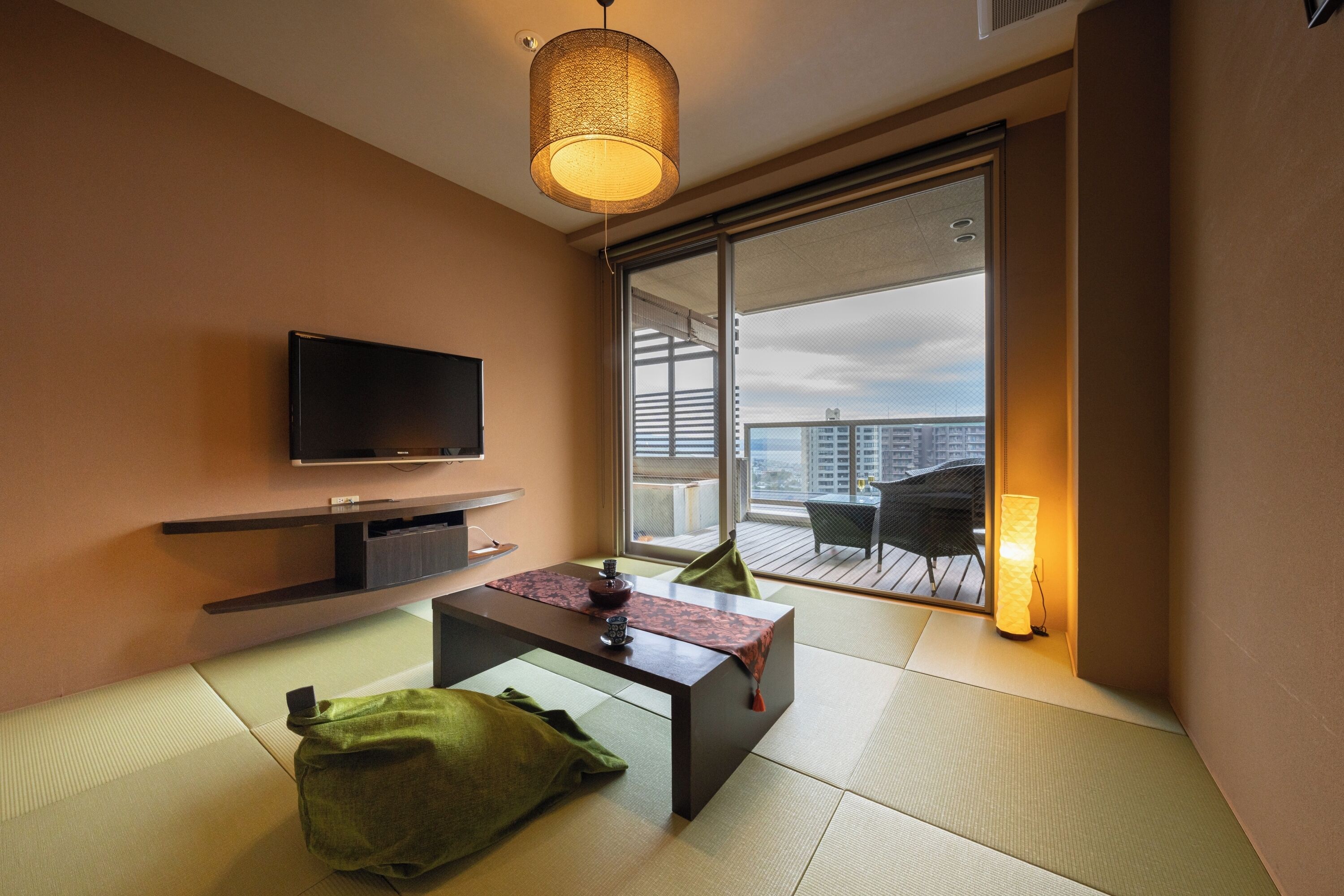 A modern living room with tatami flooring, a flat-screen TV, a low table, and a sliding glass door leading to a balcony.