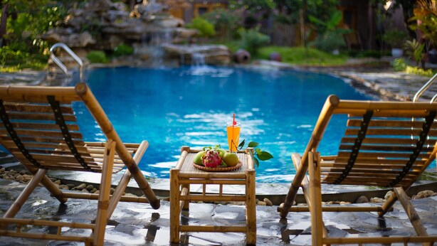 Zona de piscina con dos sillones de madera, una mesita con fruta y una bebida.