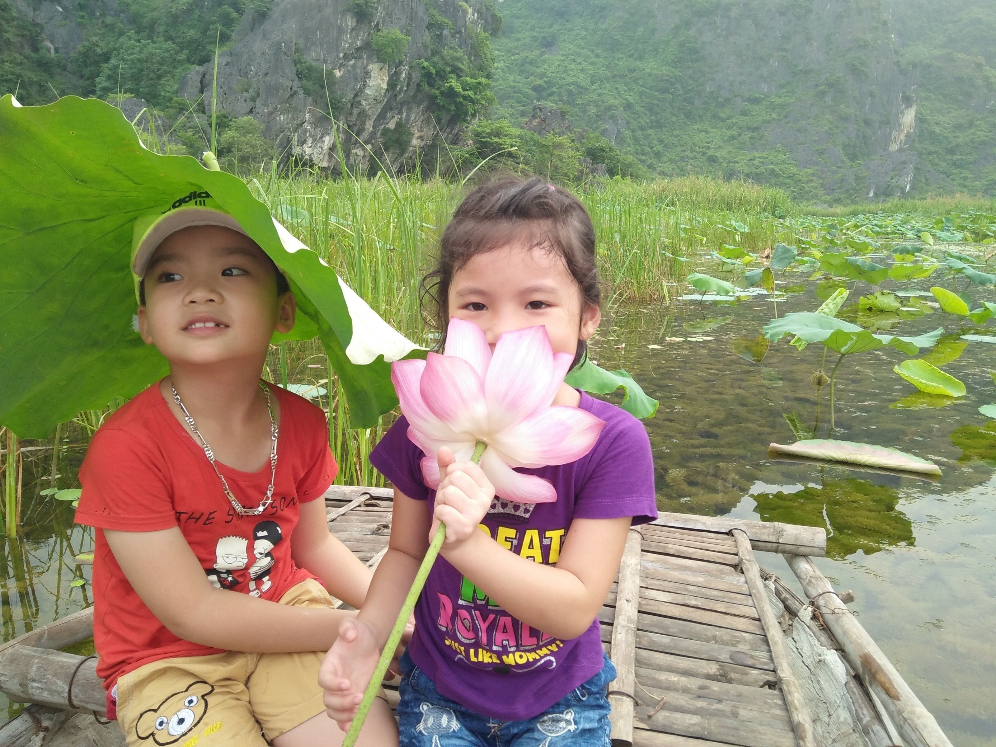 Two children sitting on a wooden dock by a pond with lily pads and greenery in the background.