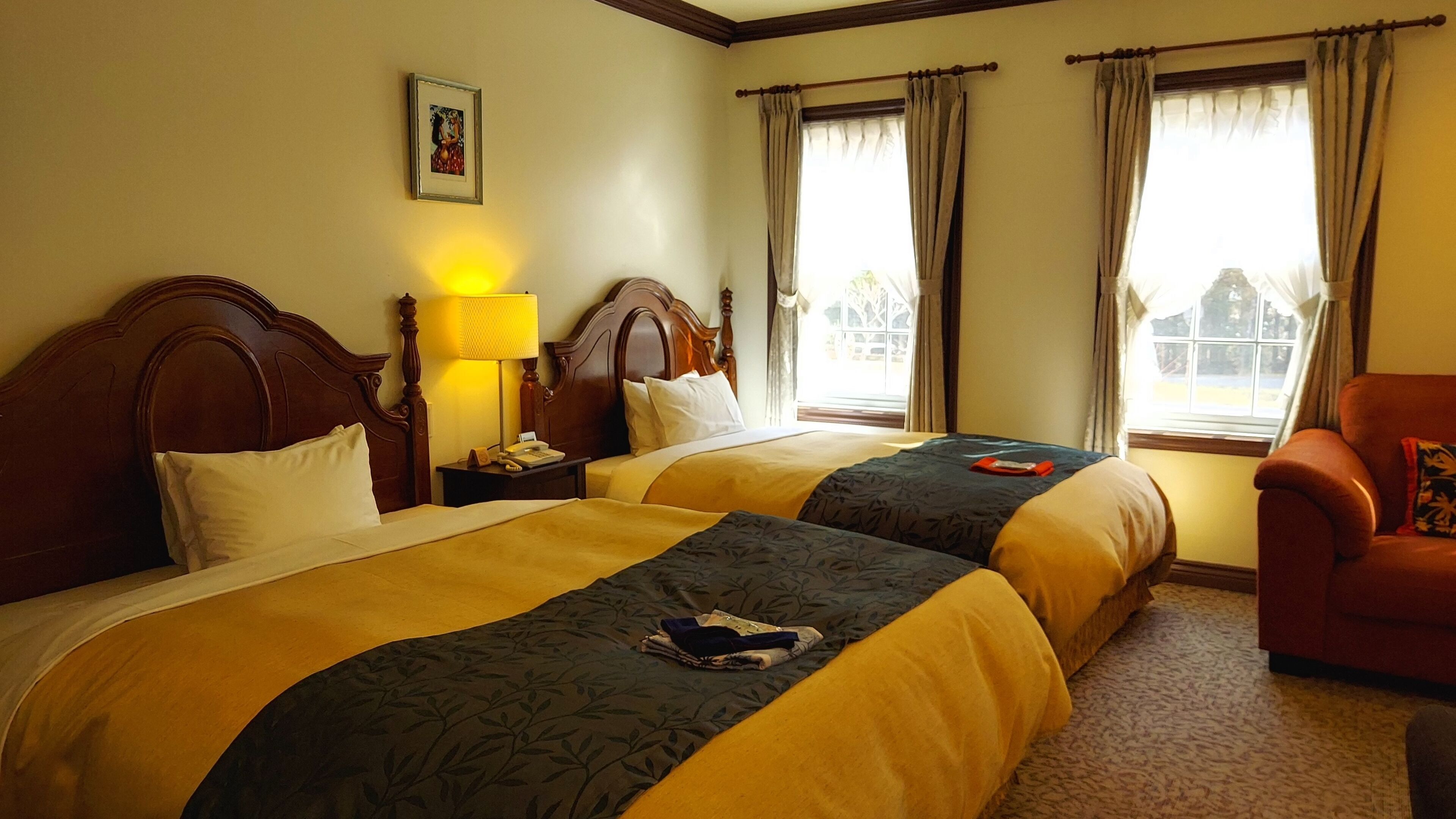A hotel room with two beds, a nightstand, a lamp, a chair, and a window with curtains.