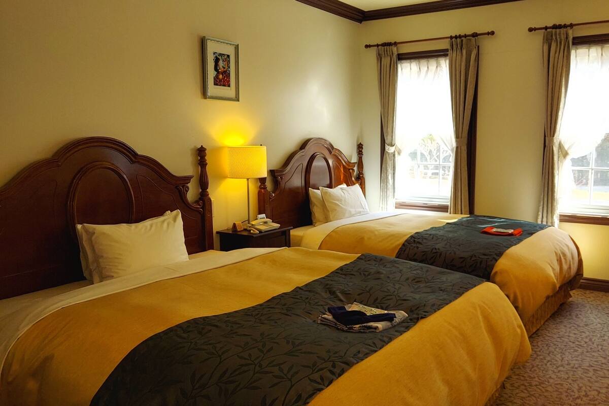 A hotel room with two beds, a nightstand, a lamp, a chair, and a window with curtains.