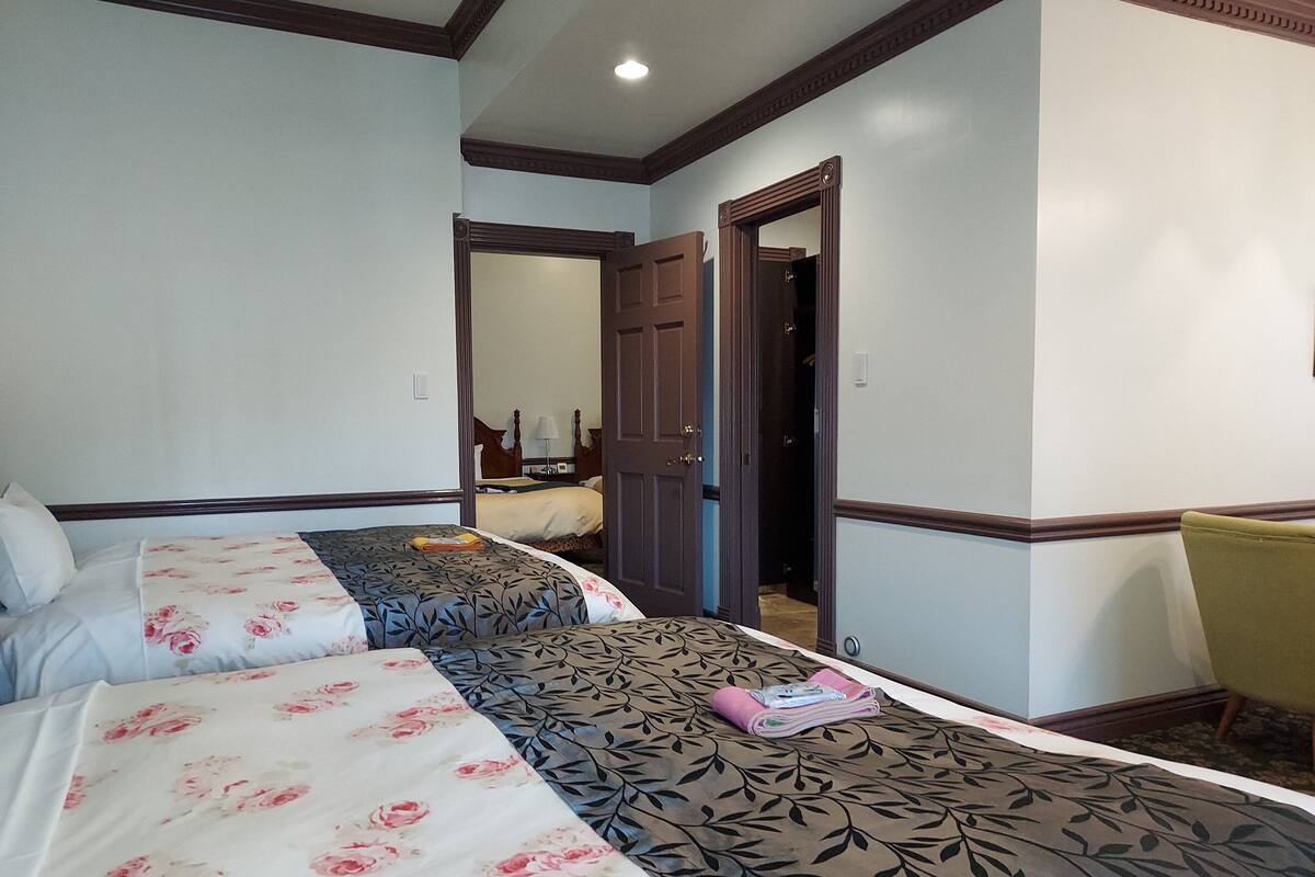 A hotel room with two beds, each with floral and leaf-patterned bedding. There is a chair and a small table with a lamp.