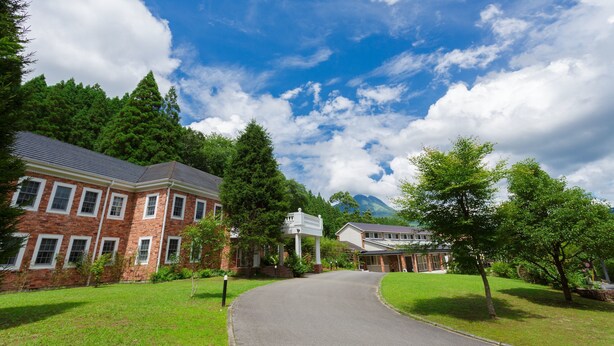 A brick building with a curved driveway, surrounded by lush greenery and trees. - Yufuin Hotel Morino Terrace (Yufu)