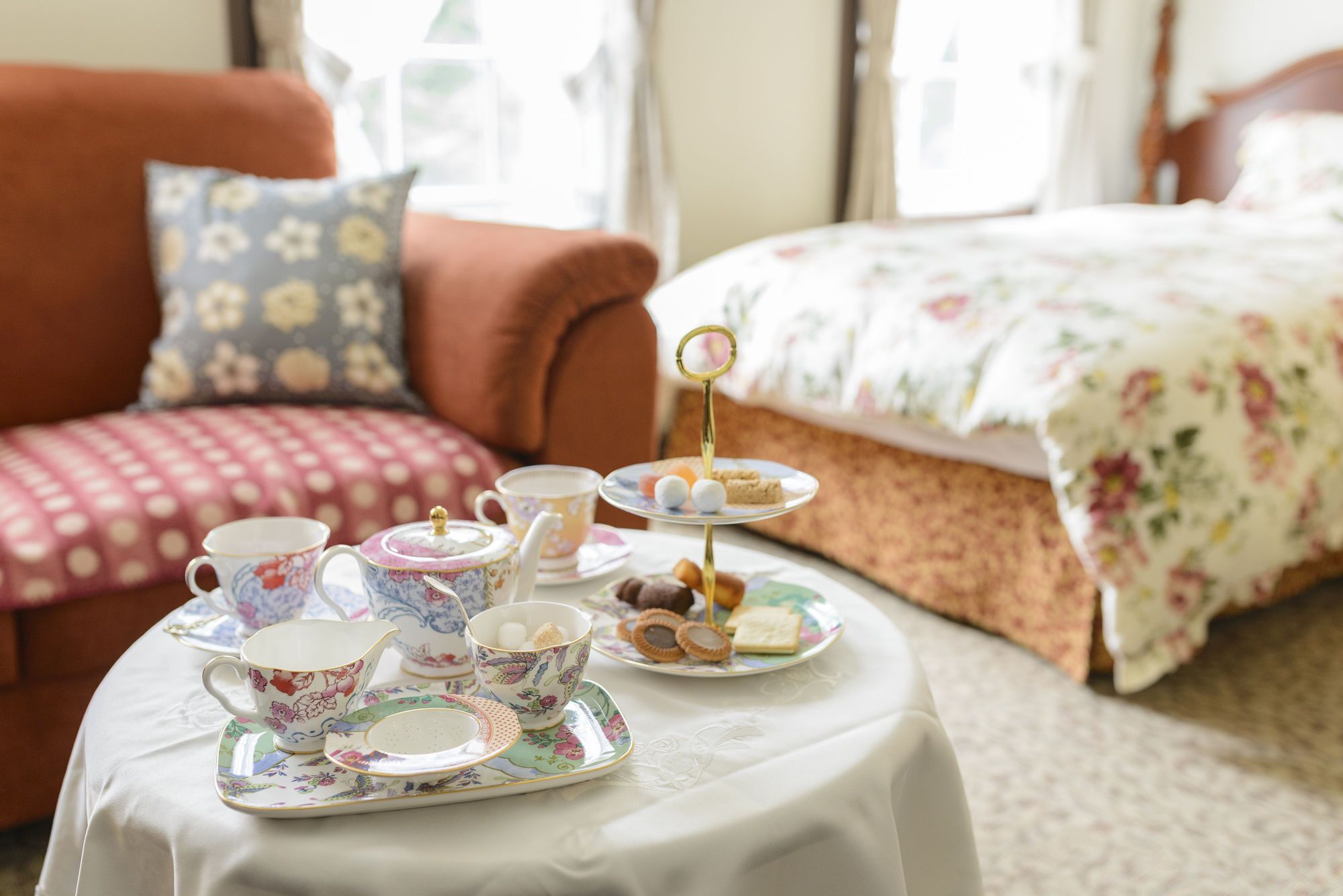 Un juego de té sobre una mesa, con una colcha estampada con flores y una silla de lunares al fondo.