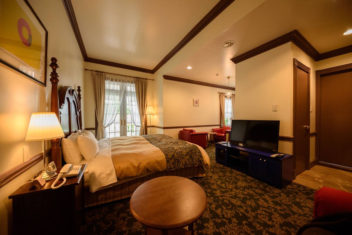 A hotel room with a bed, a desk with a lamp, a television, and a small table.