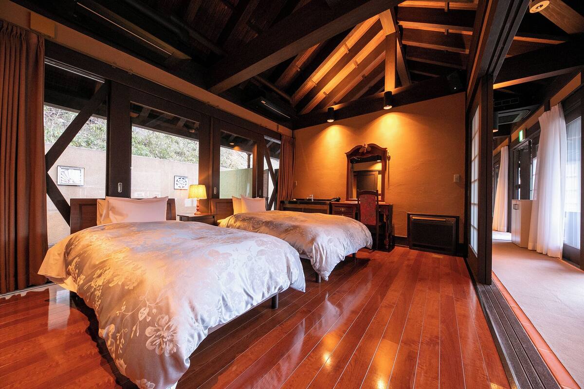 A hotel room with two beds, wooden flooring, a large mirror, and a view of the outdoors.