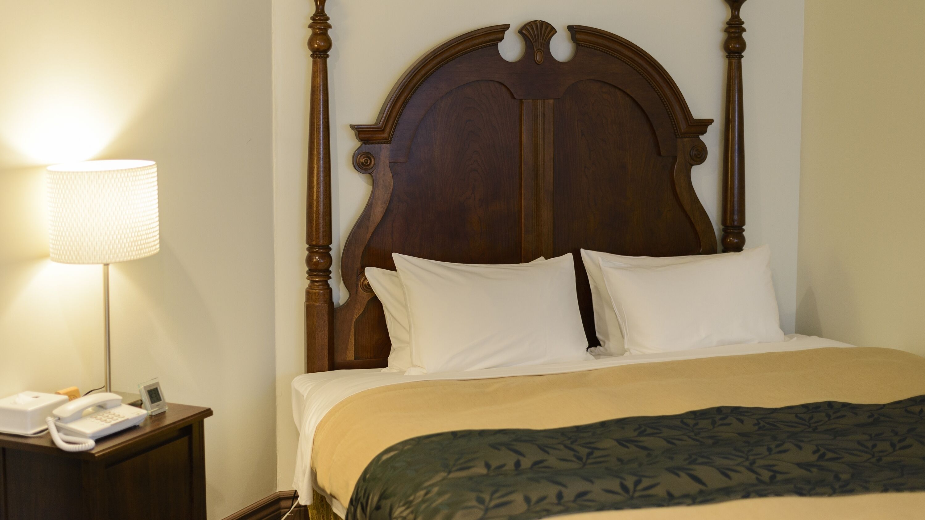 A neatly made bed with a wooden headboard, a bedside lamp, and a nightstand with a phone and a clock.